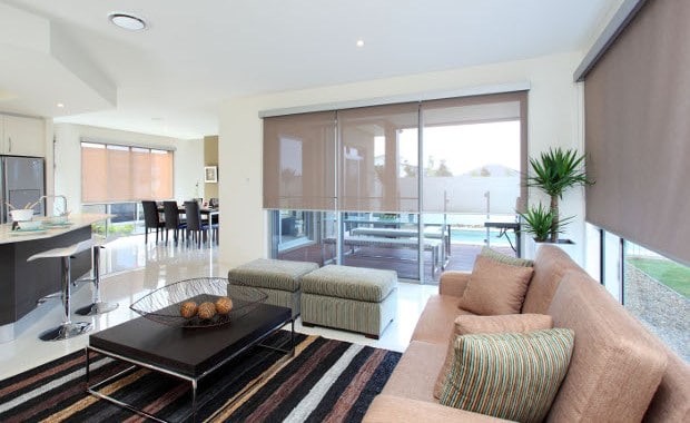 Modern living room with roller shades. Beige sofa, striped rug, and ottomans. Open floor plan with dining area and kitchen visible. Natural light.