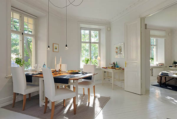 Bright dining room with natural light, white walls, and wood floors. Table set for a meal, pendant lights, and open doorway to bedroom.