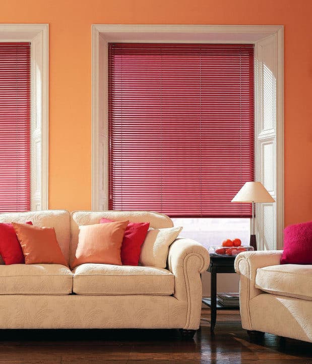 Living room with timber window coverings. The red blinds offer privacy. A sofa and chair sit on a wood floor, with a lamp on a table.