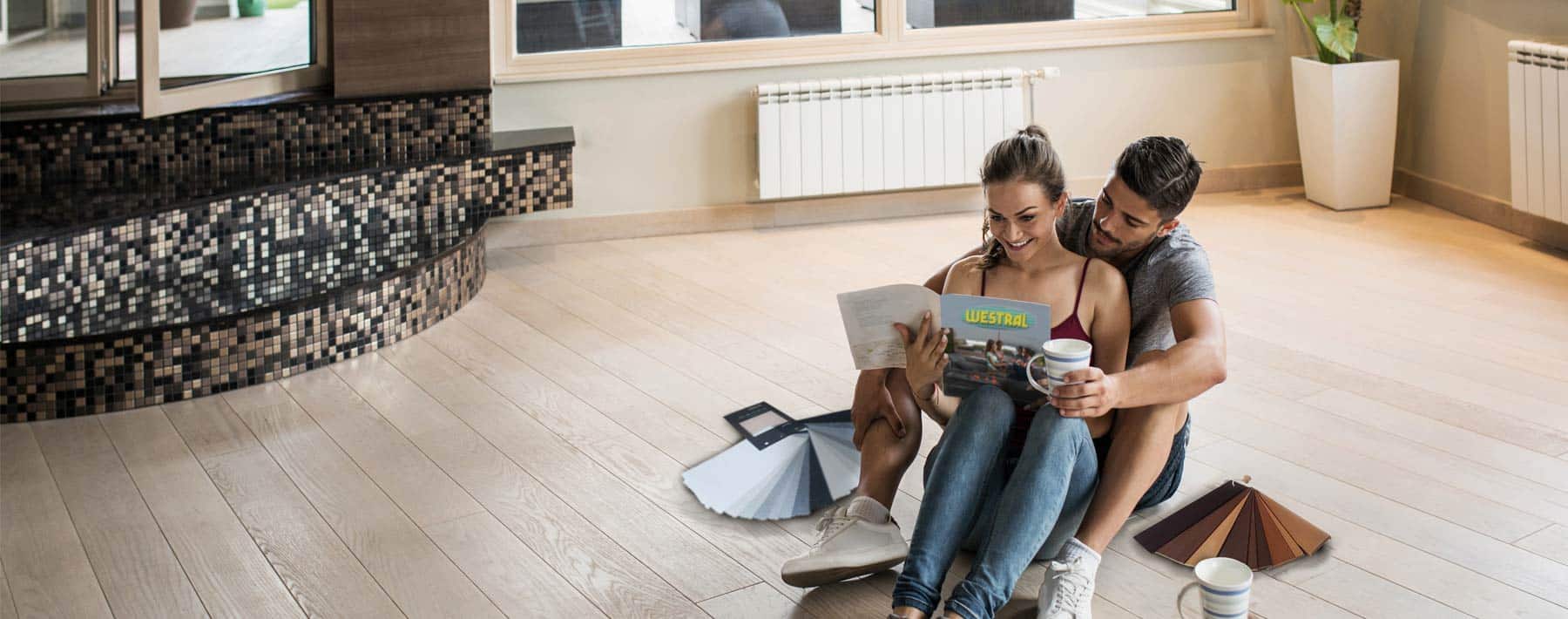 Couple sits on wood floor with paint swatches, planning home renovation. Woman holds magazine, man embraces. Radiator, mosaic tiles visible.