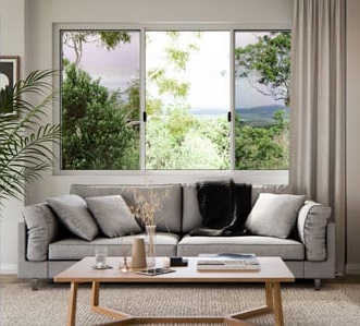 Living room interior: gray sofa with pillows, wooden table, and large window overlooking lush greenery. Natural light and neutral tones create a cozy community space.